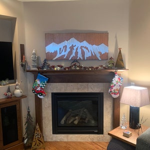 May include: A decorated fireplace mantel with Christmas stockings, a mountain-themed artwork, and festive decorations. The fireplace has a black surround and a tiled hearth. A lamp and side table are to the right.