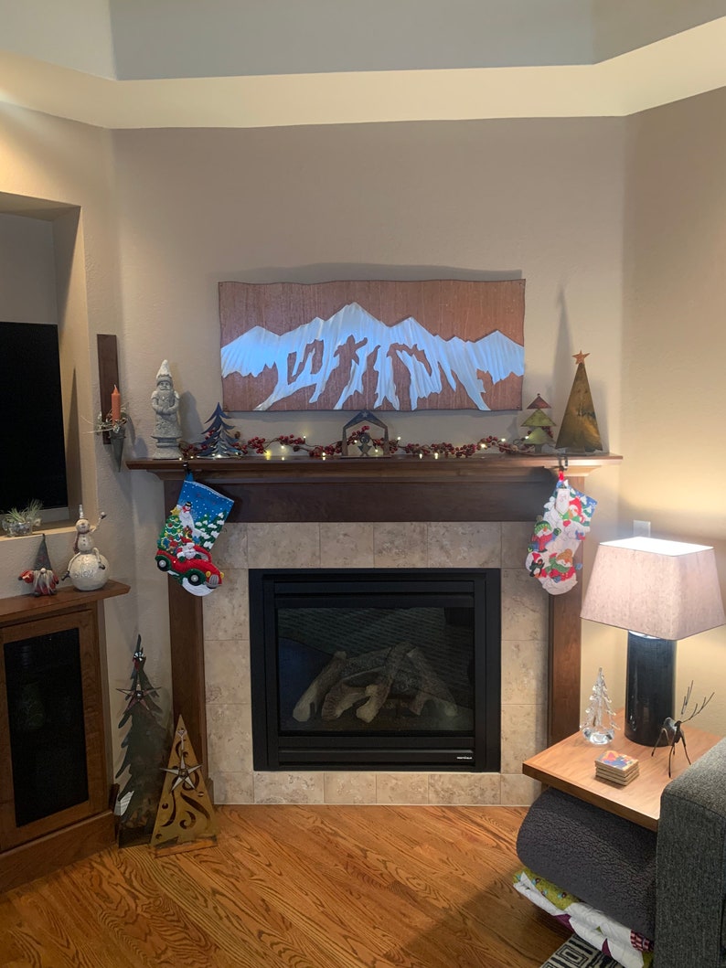 May include: A decorated fireplace mantel with Christmas stockings, a mountain-themed artwork, and festive decorations. The fireplace has a black surround and a tiled hearth. A lamp and side table are to the right.
