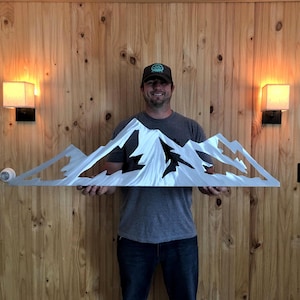 May include: A silver metal mountain range wall art piece. The artwork features a detailed cutout design of a mountain range with peaks and valleys. The piece is held up in front of a wooden wall.