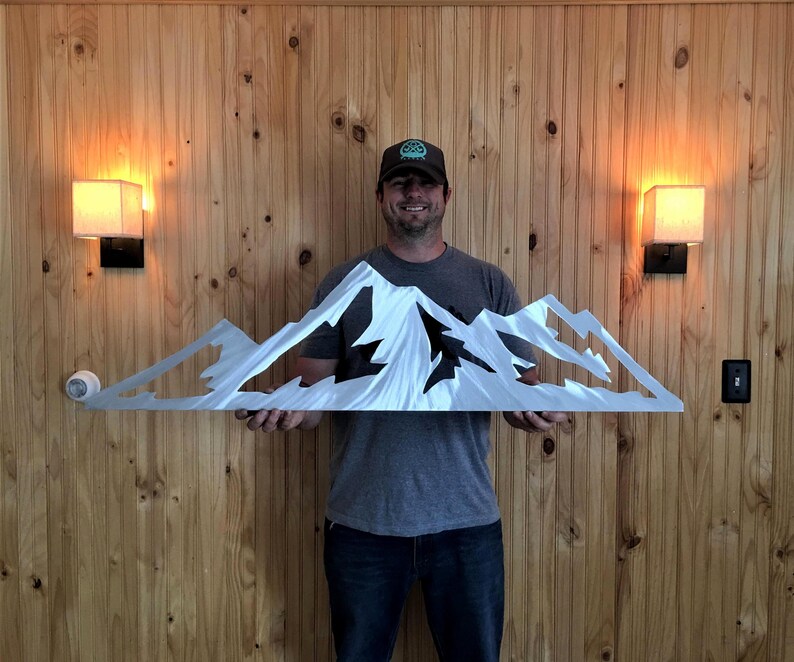 May include: A silver metal mountain range wall art piece. The artwork features a detailed cutout design of a mountain range with peaks and valleys. The piece is held up in front of a wooden wall.