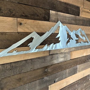 May include: A brushed metal mountain range wall art piece. The artwork features a stylized mountain silhouette with cut-out areas depicting trees. The piece is mounted on a rustic wooden plank wall, creating a contrast of textures and tones.