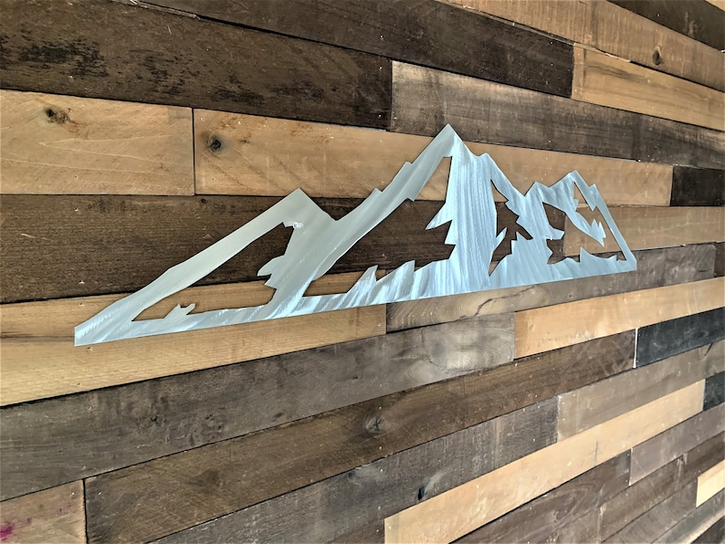 May include: A brushed metal mountain range wall art piece. The artwork features a stylized mountain silhouette with cut-out areas depicting trees. The piece is mounted on a rustic wooden plank wall, creating a contrast of textures and tones.