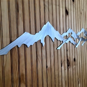 May include: A horizontal metal wall art piece featuring a stylized mountain range silhouette. The object is crafted from brushed silver-toned metal with a smooth, reflective surface, showcasing jagged peaks and valleys in a continuous, elongated design.