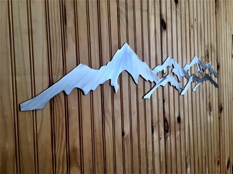 May include: A horizontal metal wall art piece featuring a stylized mountain range silhouette. The object is crafted from brushed silver-toned metal with a smooth, reflective surface, showcasing jagged peaks and valleys in a continuous, elongated design.