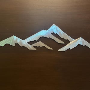 May include: A silver-toned metal wall art piece featuring a stylized mountain range silhouette. The design consists of three distinct peaks with jagged, angular edges and a brushed metallic finish that reflects light across the surface.