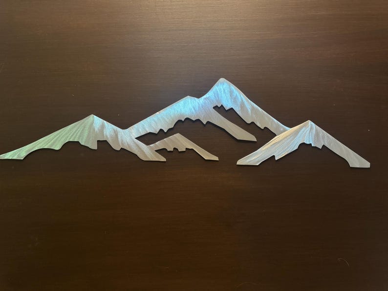 May include: A silver-toned metal wall art piece featuring a stylized mountain range silhouette. The design consists of three distinct peaks with jagged, angular edges and a brushed metallic finish that reflects light across the surface.