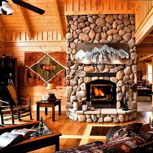 May include: A cozy cabin interior with a stone fireplace, wooden walls, and a high, sloped ceiling. A painting of a deer hangs on the wall. A lit fireplace and a ceiling fan are visible. The room is furnished with a rocking chair, a coffee table, and a lamp.
