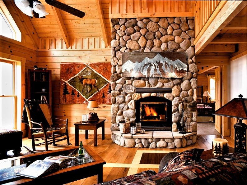 May include: A cozy cabin interior with a stone fireplace, wooden walls, and a high, sloped ceiling. A painting of a deer hangs on the wall. A lit fireplace and a ceiling fan are visible. The room is furnished with a rocking chair, a coffee table, and a lamp.