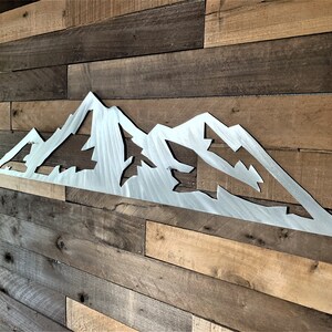 May include: A silver metal mountain range wall art piece. The artwork features a stylized mountain silhouette with cut-out shapes of trees and peaks. The piece is mounted on a wooden plank wall, creating a rustic aesthetic.