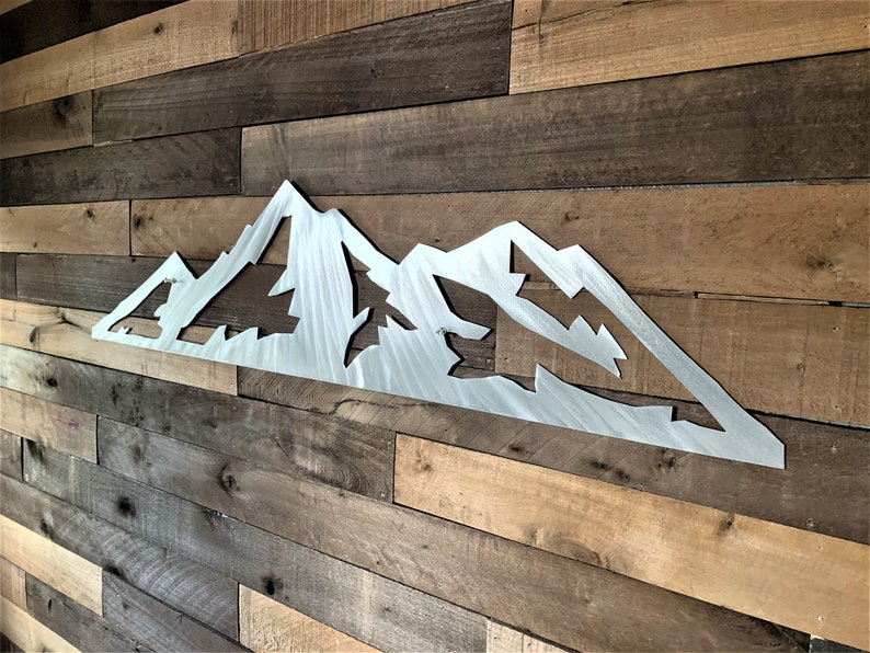 May include: A silver metal mountain range wall art piece. The artwork features a stylized mountain silhouette with cut-out shapes of trees and peaks. The piece is mounted on a wooden plank wall, creating a rustic aesthetic.