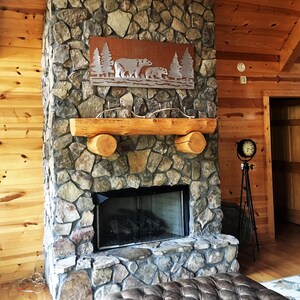 K&ouml;nnte beinhalten: Rustikaler Steinkamin mit einem h&ouml;lzernen Kaminsims und einer Metallwandkunst mit B&auml;ren und B&auml;umen. Der Kamin hat einen schwarzen Feuerraum und befindet sich in einem Raum mit Holzverkleidung.