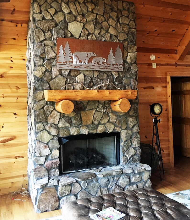 K&ouml;nnte beinhalten: Rustikaler Steinkamin mit einem h&ouml;lzernen Kaminsims und einer Metallwandkunst mit B&auml;ren und B&auml;umen. Der Kamin hat einen schwarzen Feuerraum und befindet sich in einem Raum mit Holzverkleidung.