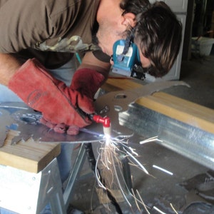 May include: A plasma cutter tool held in a red leather glove, emitting bright sparks while cutting through a flat sheet of silver-colored metal. The tool features a red handle and a white nozzle, creating a focused beam of light during the metalwork process.