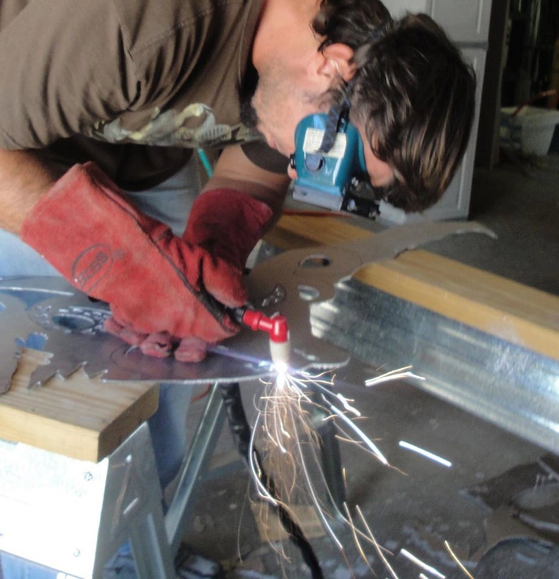 May include: A plasma cutter tool held in a red leather glove, emitting bright sparks while cutting through a flat sheet of silver-colored metal. The tool features a red handle and a white nozzle, creating a focused beam of light during the metalwork process.
