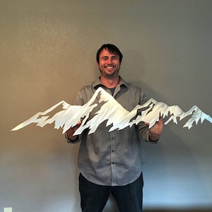 May include: A metal mountain range sculpture with a jagged, textured design. The sculpture is silver and held by a person. The artwork depicts a mountain range with peaks and valleys, suitable for home decor.