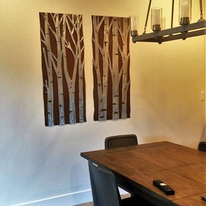 May include: Two rectangular wall art pieces with a brown background and white birch tree designs. The art is mounted on a beige wall. A dark wood dining table and black chairs are in the foreground. A chandelier hangs above the table.