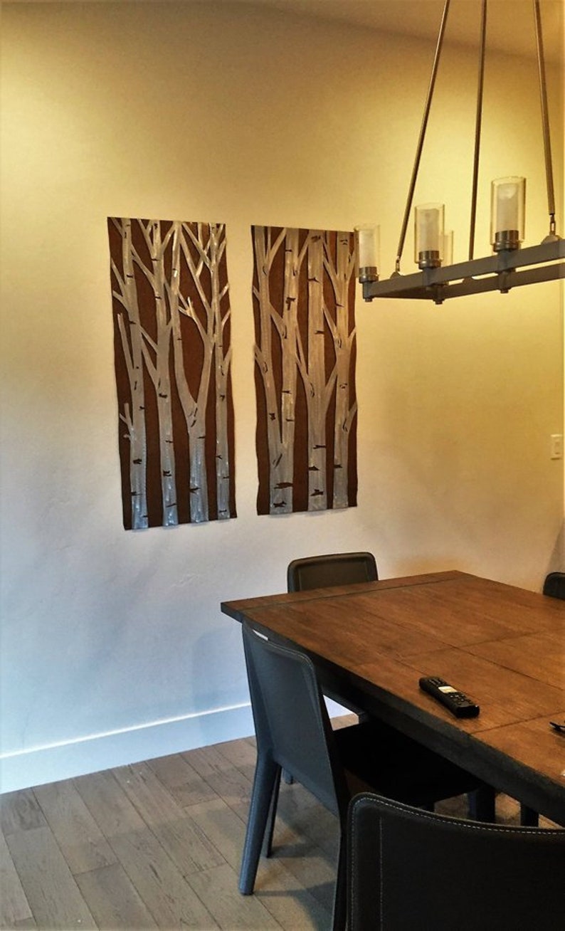 May include: Two rectangular wall art pieces with a brown background and white birch tree designs. The art is mounted on a beige wall. A dark wood dining table and black chairs are in the foreground. A chandelier hangs above the table.