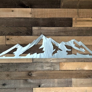 May include: A silver metal mountain range silhouette. The artwork is cut out, showing the shape of the mountains and trees. The piece is mounted on a wooden plank wall with various shades of brown.