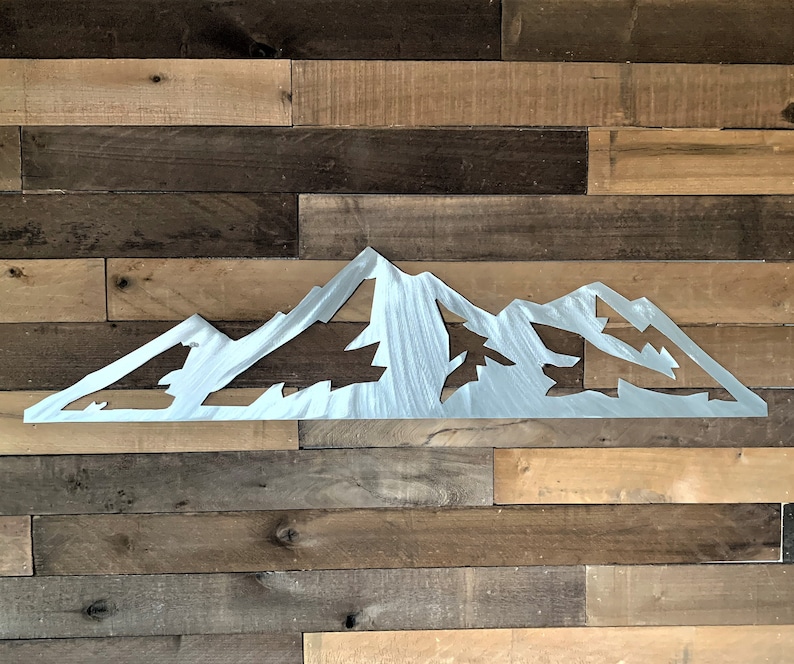 May include: A silver metal mountain range silhouette. The artwork is cut out, showing the shape of the mountains and trees. The piece is mounted on a wooden plank wall with various shades of brown.