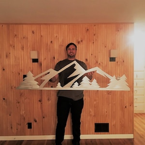 May include: A person holds a metal wall art sculpture depicting a mountain range with trees. The sculpture is white and is held in front of a wood-paneled wall.