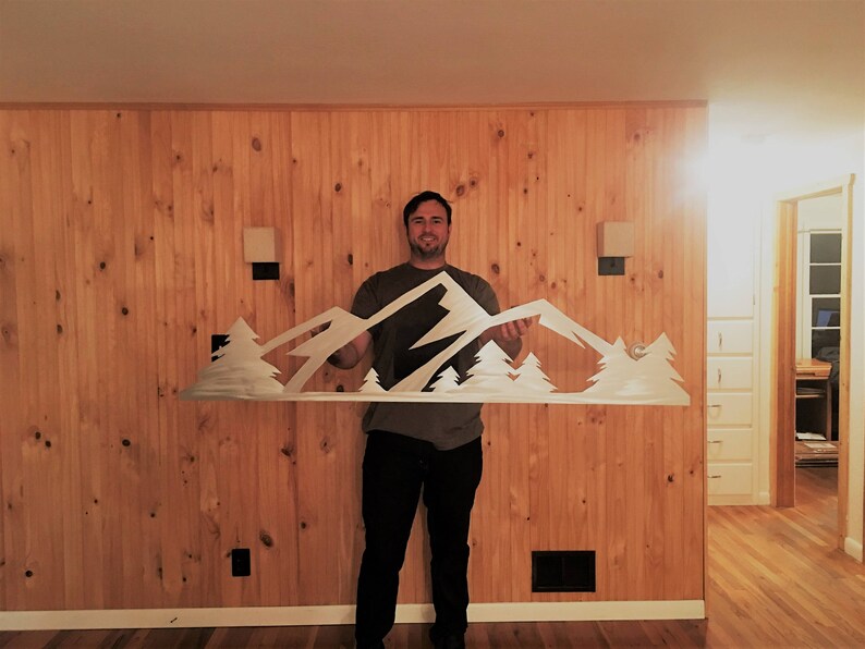 May include: A person holds a metal wall art sculpture depicting a mountain range with trees. The sculpture is white and is held in front of a wood-paneled wall.