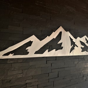 May include: A silver-colored metal mountain range wall art piece. The artwork is cut out in the shape of a mountain range with peaks and valleys. The piece is mounted on a dark, textured wall.