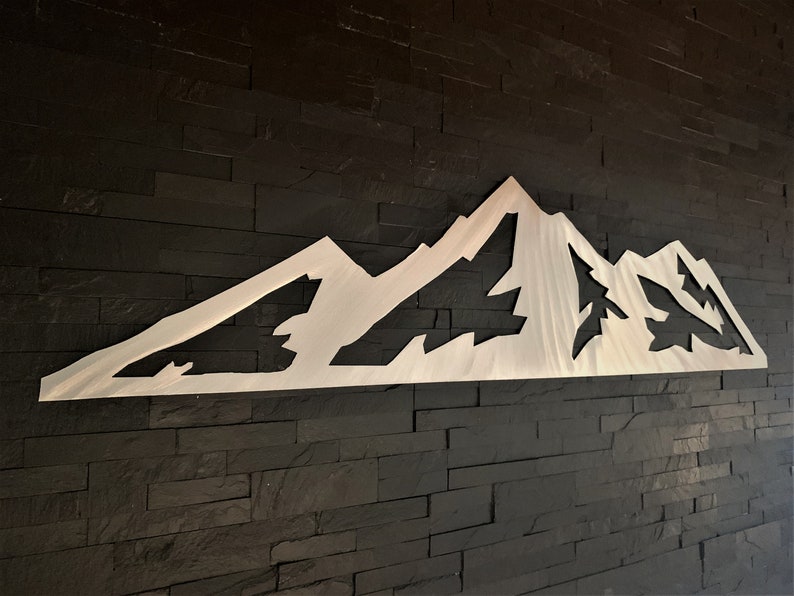 May include: A silver-colored metal mountain range wall art piece. The artwork is cut out in the shape of a mountain range with peaks and valleys. The piece is mounted on a dark, textured wall.
