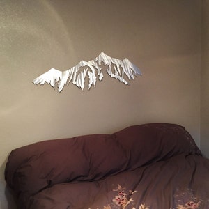 Pu&ograve; includere: Una scultura da parete in metallo di una catena montuosa in una camera da letto. La scultura &egrave; argentata ed &egrave; montata su una parete beige sopra un letto marrone con un piumino a motivi floreali.