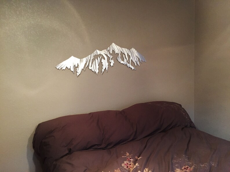 Pu&ograve; includere: Una scultura da parete in metallo di una catena montuosa in una camera da letto. La scultura &egrave; argentata ed &egrave; montata su una parete beige sopra un letto marrone con un piumino a motivi floreali.