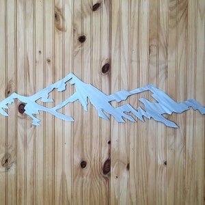 May include: A silver metal mountain range wall art silhouette. The mountain range is cut out of metal and has a brushed finish. The art is mounted on a wood panel.