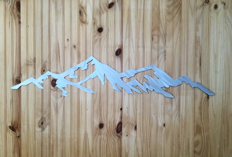 May include: A silver metal mountain range wall art silhouette. The mountain range is cut out of metal and has a brushed finish. The art is mounted on a wood panel.