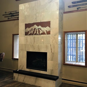 May include: A marble fireplace with a copper mountain art piece. The fireplace has a black hearth and is surrounded by windows and wooden floors. Vintage skis are mounted on the wall above.