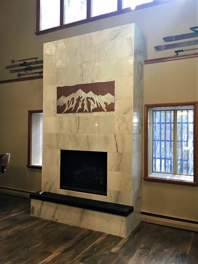 May include: A marble fireplace with a copper mountain art piece. The fireplace has a black hearth and is surrounded by windows and wooden floors. Vintage skis are mounted on the wall above.