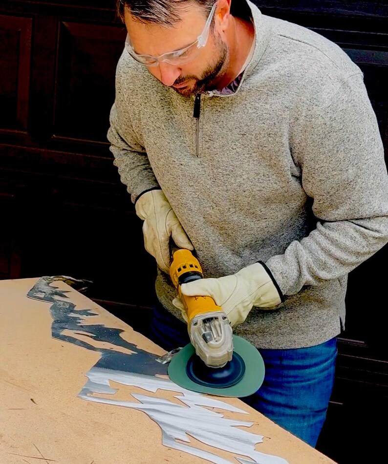 May include: A yellow power tool with a green circular sanding disc is being used to smooth the edges of a jagged, silver-colored metal sheet. The tool is held by hands wearing beige leather work gloves, while clear safety glasses are worn.