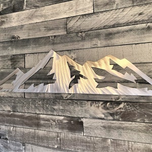 May include: A metal mountain range wall art piece. The artwork is cut from a silver-colored metal and features a detailed mountain silhouette with cut-out shapes. The piece is mounted on a wooden plank wall.