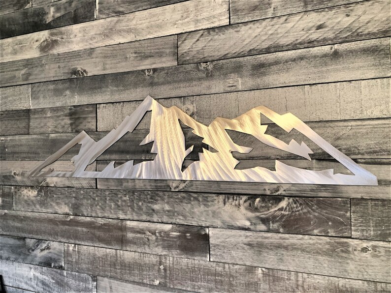 May include: A metal mountain range wall art piece. The artwork is cut from a silver-colored metal and features a detailed mountain silhouette with cut-out shapes. The piece is mounted on a wooden plank wall.
