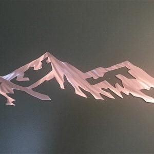 May include: A metal wall art sculpture depicting a mountain range silhouette. The sculpture is made of brushed metal and has a silver finish.