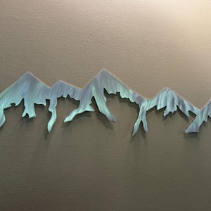 K&ouml;nnte beinhalten: Ein horizontales Wandkunstobjekt aus Metall, das die Silhouette eines Gebirgszuges darstellt. Die Oberfl&auml;che weist ein geb&uuml;rstetes Finish mit irisierenden Blau- und Gr&uuml;nt&ouml;nen auf, was den gezackten Gipfeln und H&auml;ngen eine texturierte Optik verleiht.