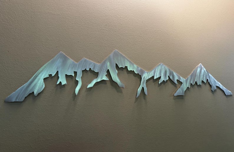 K&ouml;nnte beinhalten: Ein horizontales Wandkunstobjekt aus Metall, das die Silhouette eines Gebirgszuges darstellt. Die Oberfl&auml;che weist ein geb&uuml;rstetes Finish mit irisierenden Blau- und Gr&uuml;nt&ouml;nen auf, was den gezackten Gipfeln und H&auml;ngen eine texturierte Optik verleiht.