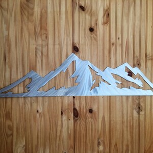 May include: A metal mountain range wall art piece. The artwork is cut from a silver-colored metal and depicts a jagged mountain range silhouette. The piece is mounted on a wooden wall with vertical planks.