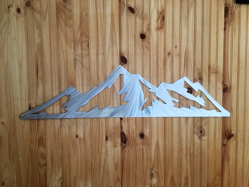May include: A metal mountain range wall art piece. The artwork is cut from a silver-colored metal and depicts a jagged mountain range silhouette. The piece is mounted on a wooden wall with vertical planks.
