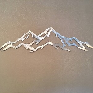 May include: A silver metal wall art sculpture depicting a mountain range with three peaks. The sculpture is cut out and has a minimalist design.
