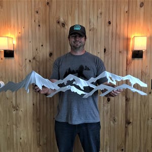 May include: A wide, white metal wall art piece featuring a layered mountain range silhouette with jagged peaks and valleys. The design consists of two distinct, overlapping horizontal sections that create a dimensional effect of snow-capped mountains.