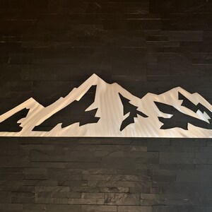 May include: A brushed metal mountain range wall art piece. The artwork features a stylized depiction of a mountain range with peaks and valleys. The metal has a silver finish and is mounted on a dark, textured wall.