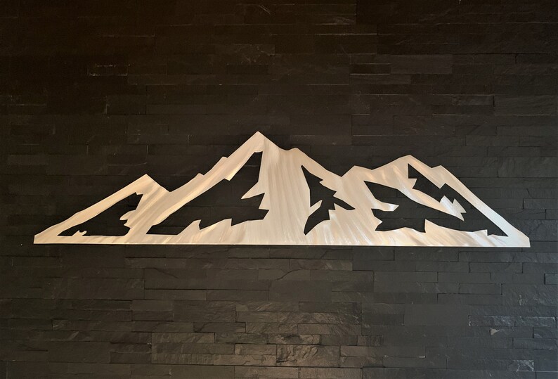 May include: A brushed metal mountain range wall art piece. The artwork features a stylized depiction of a mountain range with peaks and valleys. The metal has a silver finish and is mounted on a dark, textured wall.