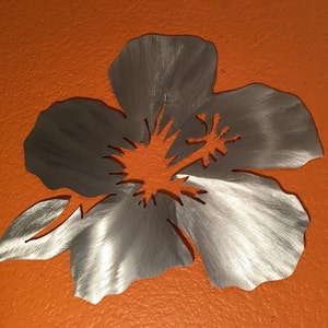 Puede incluir: Una decoraci&oacute;n de pared de metal plateado en forma de flor de hibisco. La flor est&aacute; cortada de metal y tiene un acabado cepillado. La flor est&aacute; montada sobre un fondo naranja s&oacute;lido.