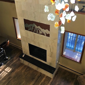 May include: A modern fireplace with a marble facade and a black hearth. Above the fireplace is a mountain-themed artwork. A decorative chandelier with glass pieces hangs from the ceiling. The room has hardwood flooring and two windows.