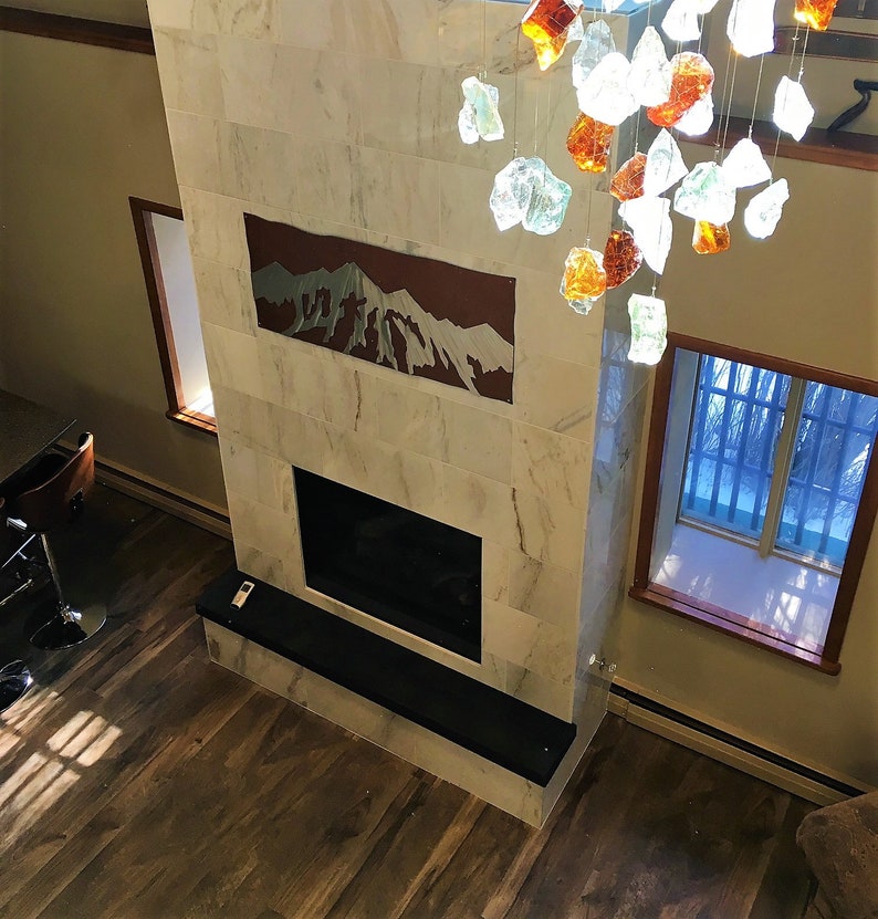 May include: A modern fireplace with a marble facade and a black hearth. Above the fireplace is a mountain-themed artwork. A decorative chandelier with glass pieces hangs from the ceiling. The room has hardwood flooring and two windows.