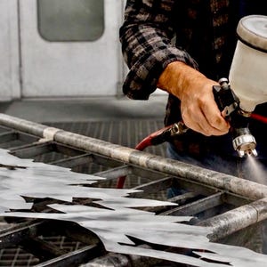 May include: A pneumatic paint spray gun with a translucent white canister is being used to apply a fine mist of paint onto a jagged, metallic, silver-colored sheet of material resting on a dark, industrial-style metal grid work surface.