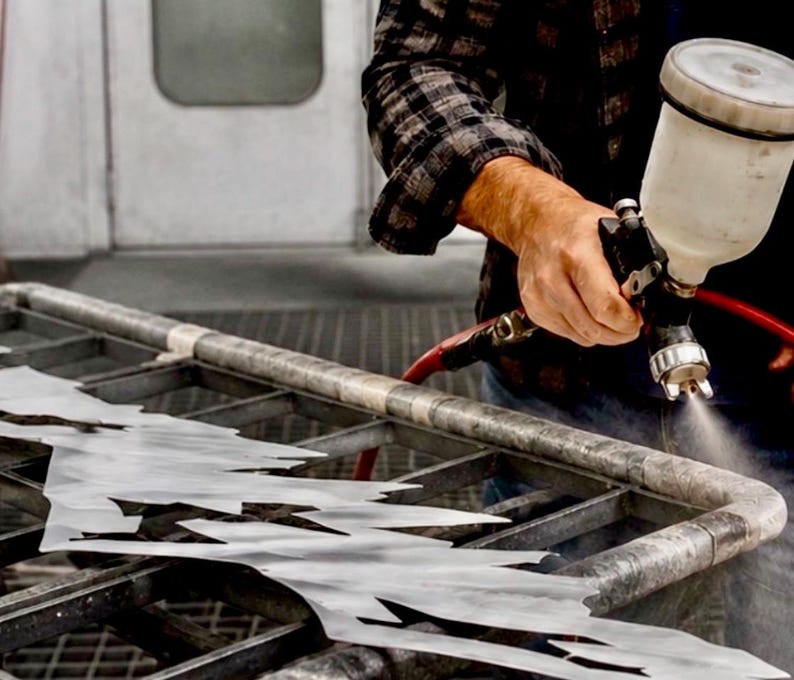 May include: A pneumatic paint spray gun with a translucent white canister is being used to apply a fine mist of paint onto a jagged, metallic, silver-colored sheet of material resting on a dark, industrial-style metal grid work surface.